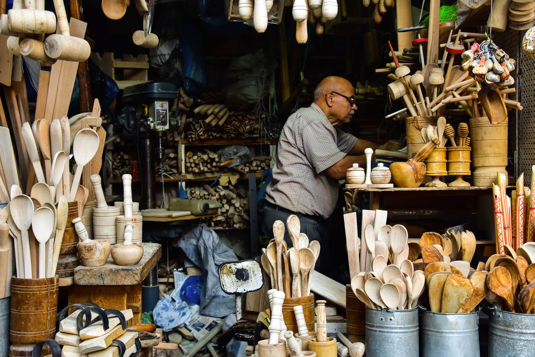 Un artisan dans son échoppe entouré de cuillères, pilons et maillets en bois exposés dans des pots et des seaux.