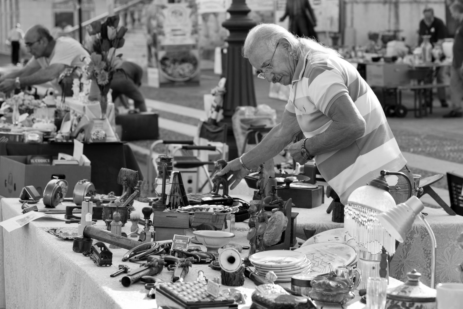 Un homme âgé se penche sur une table de brocante remplie d’objets d’occasion, outils, assiettes et lampes, dans un marché en plein air.